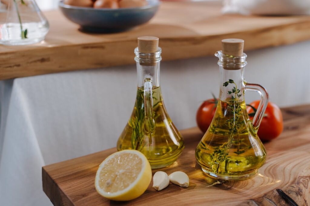 Two glass bottles of olive oil with herbs, a halved lemon, two garlic cloves, and tomatoes on a wooden surface.